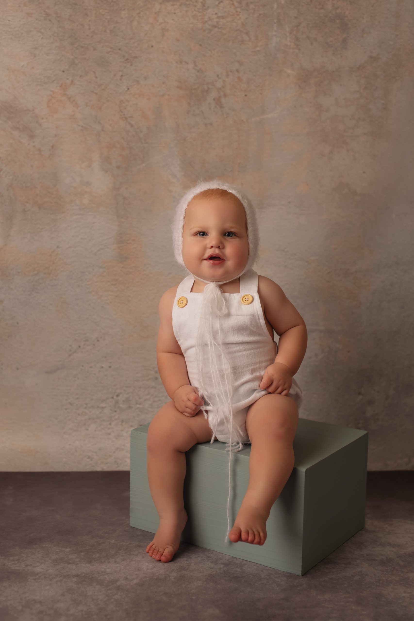 baby wearing white romper and bonnet sitting on blue box