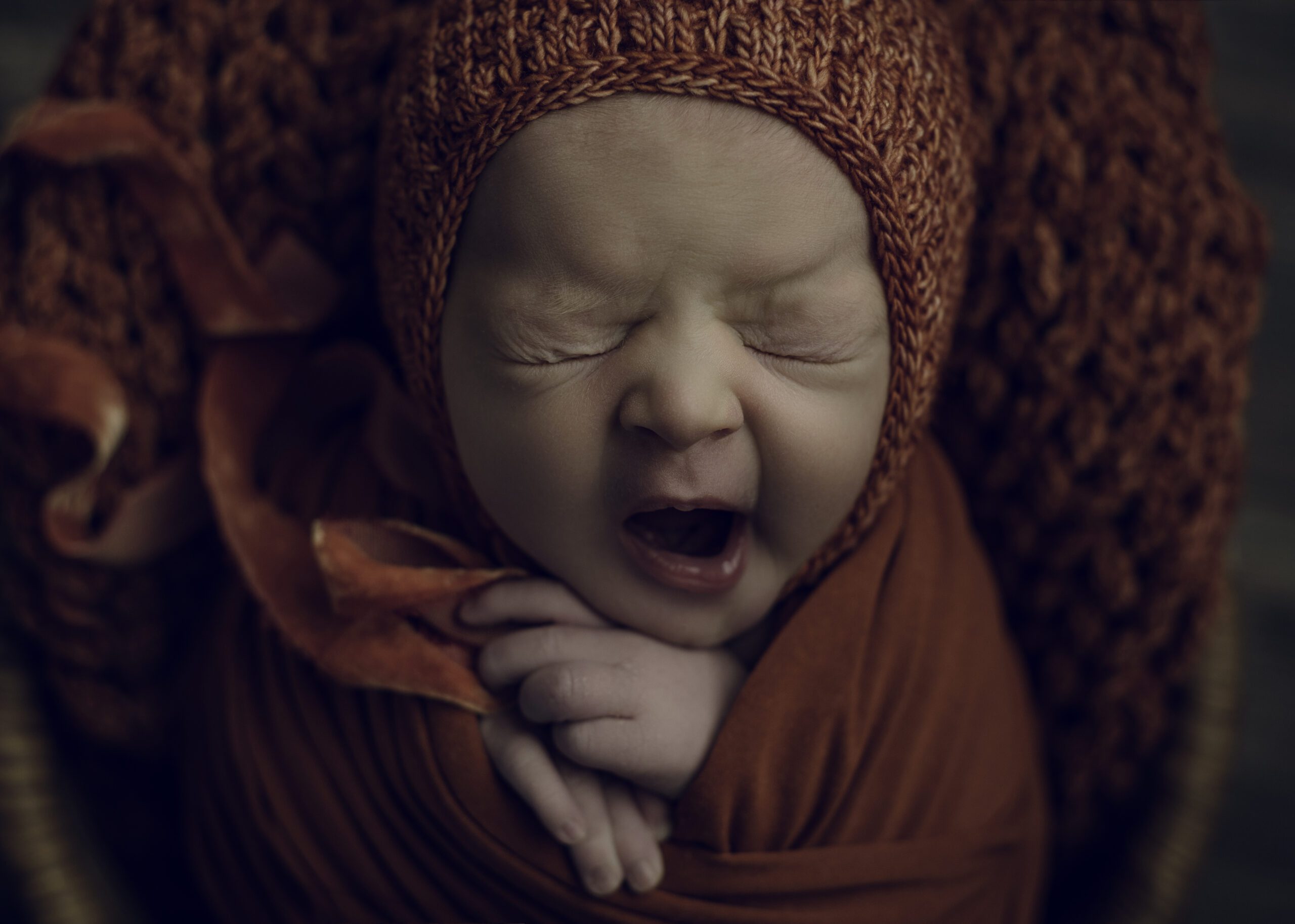 Newborn baby yawning wearing dark orange bonnet and blanket